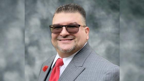 Will Torres wears a gray pinstriped suit with a red tie, a red carnation on the lapel, and sunglasses.