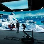 A man wearing jeans, sneakers, 和 a plaid shirt st和s in a production studio. He is facing a large LED screen showing an icy mountain range 和 a snowmobile.